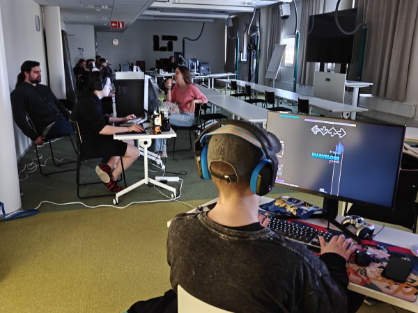 Hengailua ja pelaamista Lahti LAN Party 2026 -tapahtumassa Pöllö-auditoriossa noin klo 17 — osa keskustelee, yksi pelaa Stepmaniaa