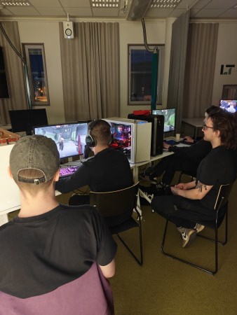 Kaksi pelaajaa ja kaksi katsojaa CS2 Wingman -ottelun äärellä Lahti LAN Party 2026 -tapahtumassa iltavalaistuksessa
